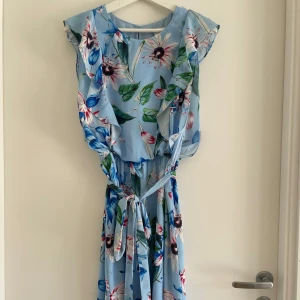 Gravidklänning/ Amningsklänning från H&M MAMA - Superfin ljusblå midiklänning  från H&M MAMA med blommigt mönster. Klänningen har volangdetaljer, knytband i midjan och är tillverkad i ett lätt, flowy material. Klänningen är avsedd som både gravidklänning och amningsklänning men går även att använda den utöver det. Superfin! 