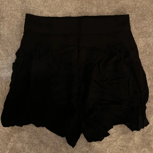 Svarta högmidjade shorts - Svarta shorts med hög midja och lös passform. De har en bred midjeresår och lätt vågiga kanter nedtill för en chill och trendig look. Perfekta för varma dagar när du vill vara både bekväm och snygg.