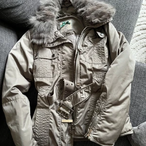 Militärgrön vinterjacka med fuskpäls  - Snygg militärgrön vinterjacka från Silvian Heach med fuskpäls, bälte i midjan och fickor framtill. Jackan har dragkedja och knappar 🧚🏽🌸