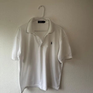 Vit pikétröja från Ralph Lauren - Säljer en klassisk vit pikétröja från Ralph Lauren med korta ärmar och den ikoniska loggan broderad på bröstet. Perfekt för en stilren och avslappnad look. Passar till både jeans och chinos. Hör av dig vid intresse!