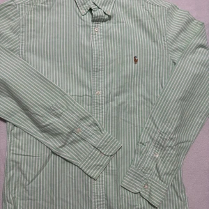 Grönrandig skjorta Polo Ralph Lauren - Snygg grön och vit randig skjorta från Polo Ralph Lauren med klassisk krage och broderad logga på bröstet. Skjortan har lång ärm, knappar framtill och är i bomull. Perfekt för dig som gillar stilrena och fräscha plagg.