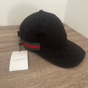 Svart keps från Gucci med GG-mönster - Snygg svart keps från Gucci med klassiskt GG-monogram över hela kepsen. Har en grön och röd rand på sidan och justerbar rem baktill i skinn. Perfekt accessoar för att lyfta din outfit med en lyxig touch.