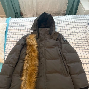 Canada Goose Wyndham grå black label  - Säljer en svart dunjacka från Canada Goose med huva och avtagbar päls. Jackan har klassisk patch på ärmen, dragkedja och knappar framtill samt stora fickor. Perfekt för kalla vinterdagar och riktigt snygg streetstil.