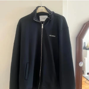 Svart bomberjacka från Les Deux - Mörk blå zip tröja från Les Deux med stilren design och broderad logga på bröstet. Jackan har hög krage, hel dragkedja framtill och ribbade muddar. Perfekt för dig som gillar en clean och sportig look.