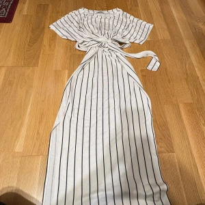 Vit långklänning med svarta ränder Bershka - Supersnygg långklänning från Bershka i vitt med svarta vertikala ränder. Klänningen har korta ärmar, knytband i midjan och en avslappnad passform. Perfekt för dig som gillar en stilren och trendig look. Har dock en lite fläck