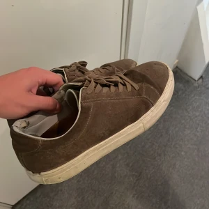 Bruna mockasneakers från Vagabond - Säljer ett par bruna sneakers från Vagabond i mocka med vita, platta sulor och klassisk snörning. Skorna har en stilren design med rund tå och diskreta sömmar. Perfekta för dig som gillar en enkel men snygg look.