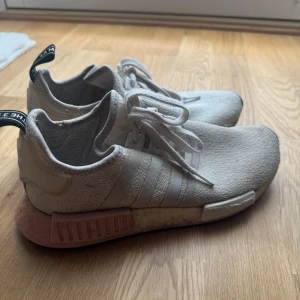 Adidas NMD R1 sneakers i vit och rosa - Adidas NMD R1 sneakers i vit mesh med klassiska tre ränder på sidan och rosa detaljer på sulan. Skorna har svart och vit dragflik bak, snörning och en bekväm, dämpad sula. Perfekta för dig som gillar streetstyle och vill ha en snygg och sportig look.