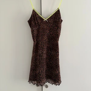 Leopardmönstrad brun kortklänning - Snygg kort klänning med leopardmönster i brunt och svart. Tunna neon-gula axelband och spetsdetalj vid v-ringad hals, samt en liten rosett framtill. Klänningen är lined och har en vågig kant nedtill.
