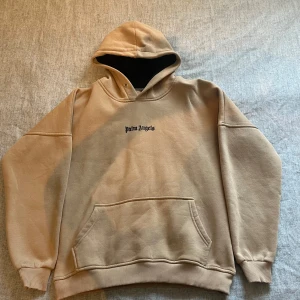 Beige hoodie från Palm Angels - Säljer en beige hoodie från Palm Angels med svart logga på bröstet och stor text på ryggen. Perfekt för dig som gillar streetwear och vill sticka ut med en snygg och clean look. Vid snabb affär kan man få plagget för ett billigare pris!