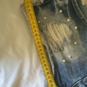 Blå jeansshorts med spets och pärlor - Snygga blå jeansshorts med ljus tvätt, med vit spets och små pärlor på sidorna. Shortsen har slitna detaljer vilket verkligen är jättesnyggt. Säljer pågrund av att de är för små för mig. De är ganska små i storleken men passar xs-s storlek.  Midjemått 37cm❤️Dålig bild men där låren sitter är måtten 24-24 cm ungefär❤️