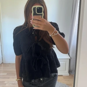 Svart blus med knyt och puffärm - Säljer en svart blus med puffiga korta ärmar och tre snygga knyt framtill. Blusen har en cool, skrynklig textur och en lite lös passform som ger en chill vibe. Perfekt att styla med jeans eller kjol för en trendig look.