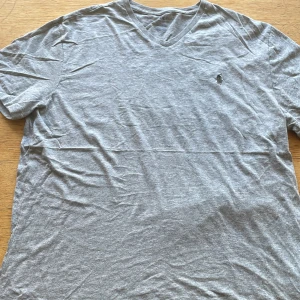 Grå v-ringad t-shirt från Polo Ralph Lauren - Snygg grå t-shirt från Polo Ralph Lauren med v-ringad hals och klassisk broderad logga på bröstet. T-shirten är kortärmad och gjord i mjuk bomull, perfekt för chill dagar. Enkel och stilren design som funkar till jeans eller shorts.