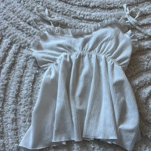 Vit volangig linnetopp med knytband - Supersöt vit linnetopp med volangkant upptill och smala axelband som knyts. Toppen har en luftig och flowy passform med rynkade detaljer över bysten. Perfekt för varma dagar och ger en romantisk vibe.