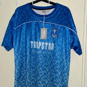 Blå Trapstar fotbollströja med tryck - Snygg blå fotbollströja från Trapstar med vit logga och text på bröstet, samt nummer 22 på ryggen. Tröjan har ett randigt mönster i olika blå nyanser, broderad emblem med tre stjärnor och vita detaljer. Tillverkad i lätt och ventilerande polyester.