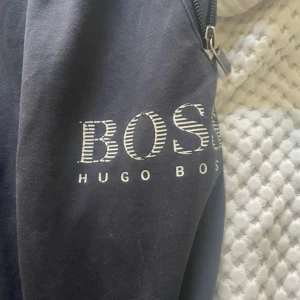 Svarta mjukisbyxor från Hugo Boss - Svarta mjukisbyxor från Hugo Boss med vit logga på benet. Byxorna har resår i midjan med snörning och dragkedjeficka på sidan. Tillverkade i mjukt bomullsmaterial, perfekta för chill eller träning. Snygg och enkel design med smal passform.                                                                          Obs! Kommer med defekter (se bild)                    För övrigt cond 6,5/10 (går att sy ihop)