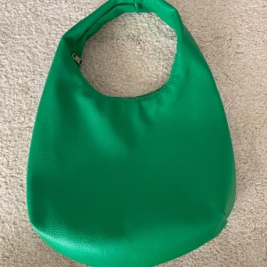 Grön axelväska i skinnimitation som ny - Säljer en snygg grön axelväska i skinnimitation med rundad form och bred axelrem. Väskan har en silvrig dragkedja upptill och ett innerfack för småprylar. Perfekt storlek för att få plats med allt du behöver till vardagen. Ser ut som ny.💚💚