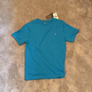 Blå t-shirt från Polo Ralph Lauren - Snygg blå t-shirt från Polo Ralph Lauren med klassisk rund hals och korta ärmar. T-shirten har en liten gul broderad logga på bröstet och är gjord i mjuk bomull som känns skön mot huden. Perfekt för dig som gillar stilrena och ikoniska plagg.