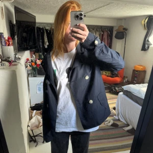 Mörkblå skepparkavaj från Broadway - Snygg mörkblå skepparkavaj från Broadway med dubbelknäppning och stora ljusa knappar. Jackan har klassisk krage, två fickor framtill och är helfodrad med randigt innerfoder. Perfekt för dig som vill ha en stilren och tidlös look.