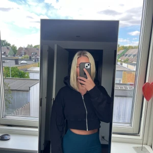 Svart croppad hoodie med dragkedja - Svart croppad hoodie med dragkedja framtill och huva. Tröjan har långa ärmar och en loose passform som ger en avslappnad vibe. Perfekt för lager-på-lager och funkar året runt. Enkel och stilren design utan tryck eller mönster.