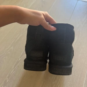 Svarta UGG boots i mocka - Säljer ett par klassiska svarta UGG boots med rund tå och platt sula. Skorna är tillverkade i mjuk mocka och har en fluffig insida som håller fötterna varma. Perfekt för dig som vill ha en chill och cozy stil under höst och vinter. Pris kan diskuteras 