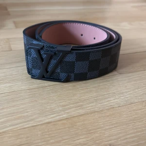 Bälte - Snygg bälte med klassiskt Damier-mönster i svart och grått. Bältet har en stor logga som spänne och är gjort i skinn med en lyxig känsla. Perfekt accessoar för att lyfta din outfit. Tveka inte för att ställa frågor.