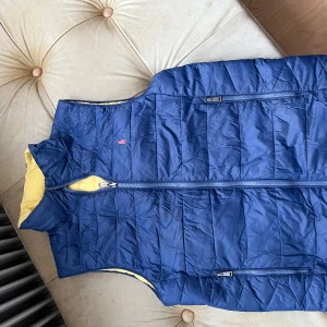 Blå quiltad dunväst från Polo Ralph Lauren - Quiltad dunväst från Polo Ralph Lauren i marinblått med gul insida. Västen har dragkedja framtill, två sidofickor med zip och broderad logga på bröstet. Perfekt för lager-på-lager och chill stil. Materialet är polyester och dun, riktigt lätt och skön.