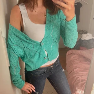 Turkos stickad kofta från Molly Bracken - Snygg turkos stickad kofta från Molly Bracken med genombrutet mönster och knappar framtill. Croppad modell med lång ärm och v-ringad hals. Perfekt att slänga över ett linne för en chill och trendig look.