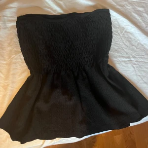 Svart bandeau tubtopp från Gina Tricot - Supersnygg svart bandeau tubtopp från Gina Tricot med smockad överdel och utsvängd nederdel. Toppen är ärmlös och axelbandslös, perfekt för att skapa en cool och trendig look. Mjuk och stretchig känsla som sitter bekvämt på plats. Kan också användas som kjol. Aldrig använd! HELT NY