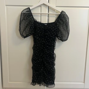 Svart prickig kortklänning med puffärm - Supersöt svart kortklänning med vita prickar och transparenta puffärmar. Klänningen har rynkade detaljer längs hela kroppen och en figurnära passform. Perfekt för dig som vill sticka ut med en lekfull och trendig look.