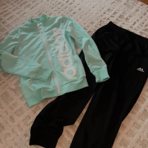 Svarta Adidas mjukisbyxor och zip-up set - Svarta mjukisbyxor från Adidas med klassisk logga på låret. Byxorna har resår i midjan och vid bensluten för en skön och avslappnad passform. Perfekta för chill eller träning, tillverkade i mjukt material. Tillsammans med en turkos zip-up med klassiska logot
