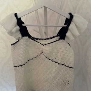 Vit stickad blus med volang - Supersöt vit stickad blus med svarta detaljer och volang på axelbanden. Blusen har en lätt rynkad effekt framtill och är perfekt för dig som gillar en romantisk och unik stil. Luftigt stickat material som passar till många outfits.