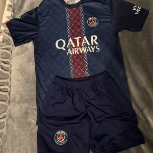 PSG matchställ blå Qatar Airways - Snyggt PSG matchställ i mörkblått med röd och vit rand på tröjan, Qatar Airways-tryck och klubbmärke på både tröja och shorts. Kortärmad tröja i lätt och sportigt material, perfekt för fotbollsträning eller att bära som supporter.