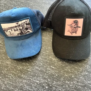 Två schyssta kepsar med tryck - Säljer två kepsar med mesh baktill. Ena kepsen är blå med ett svartvitt strandmotiv framtill, den andra är svart med ett tryck av ett djur med keps och kniv. Båda har böjd skärm och justerbar passform. Perfekt för dig som vill sticka ut med coola prints.