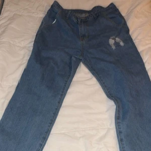 Blå jeans med stora vingar bak - Unika blå jeans med stora silvriga vingar tryckta på baksidan och mindre vingar framtill. Raka ben och klassisk passform. Perfekta för dig som vill sticka ut med coola detaljer. Materialet är klassisk denim.