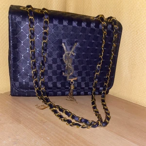 Svart YSL axelväska med kedja - Snygg svart axelväska från YSL med rutigt mönster och guldiga detaljer. Väskan har en lång kedjerem i guld och svart, samt YSL-logga framtill med hängande tofs. Perfekt storlek för det viktigaste och ger en lyxig känsla till din outfit.