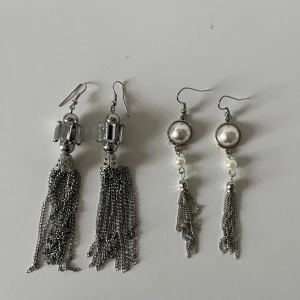 Två par silvriga örhängen  - Säljer två par coola örhängen i silverfärg. Ena paret har glittriga stenar och långa kedjor, det andra har stora pärlor och kedjor som hänger ner.