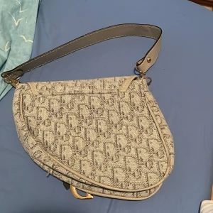 en väska i beige och grått - Säljer en ikonisk  med beige och grått monogrammönster, bred axelrem i skinn och snygga gulddetaljer. Väskan har den klassiska sadelformen och ett stort D-spänne framtill. Perfekt statement piece för dig som vill sticka ut.