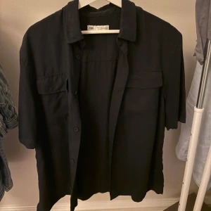 Blå skjorta från Zara - Stilren svart overshirt från Zara i storlek M Skjortan har korta ärmar, klassisk krage, knäppning framtill och två bröstfickor med lock. Perfekt att bära öppen över en t-shirt för en avslappnad look.