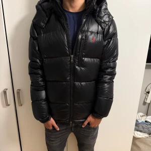 Pufferjacket från polo Ralph Lauren  - Säljer en svart pufferjacka från Polo Ralph Lauren i storlek M. Jackan har glansig finish, huva, dragkedja framtill och en röd logga på bröstet. Nyskick inga deffekter som ny, nypris-6000 mitt pris 3500, pris kan diskuteras vid snabba affärer 