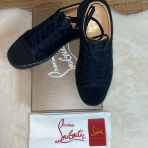 Mörkblå sneakers från Christian Louboutin - Snygga mörkblå sneakers från Christian Louboutin i mocka med klassisk röd sula och broderad logga baktill. Skorna har rund tå, platt sula och snörning. Insidan är i beige skinn för extra lyxig känsla. Perfekta för dig som vill sticka ut med stil.