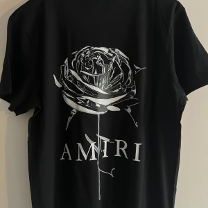 Svart Amiri t-shirt med tryck - Svart t-shirt från Amiri i mjuk bomull med coolt tryck fram och bak. Framsidan har Amiri-loggan med färgstänk och baksidan har en stor silverfärgad ros och Amiri-tryck. Klassisk passform och rund hals, perfekt för en edgy streetstyle.