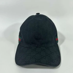 Svart keps från Gucci med GG-mönster - Exklusiv svart keps från Gucci med diskret GG-monogram över hela tyget. Kepsen har klassisk böjd skärm och ikoniska gröna och röda ränder på sidan. Tillverkad i bomull och har justerbar rem bak i skinn. Perfekt för dig som vill ha en stilren och lyxig accessoar.