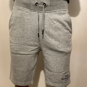 Grå mjukisshorts Peak Performance - Snygga grå mjukisshorts från Peak Performance med broderad logga på benet. De har hög midja, snörning i midjan och fickor både fram och bak. Materialet är mjukt och skönt, perfekt för chill eller träning. Klassisk loose passform och stilren look.