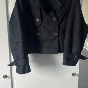 Svart kort trenchcoat från H&M - Endast använd en gång .Kort svart trenchcoat från H&M med dubbelknäppning, stora slag och bältesdetaljer vid ärmsluten. Jackan har en clean och stilren look, perfekt för dig som gillar minimalistisk stil. Tillverkad i ett slätt tyg som känns lätt men ändå strukturerat.