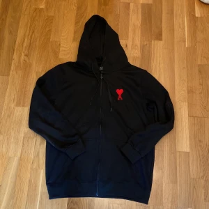 Svart hoodie från Ami med rött hjärta - Svart hoodie från Ami Alexandre Mattiussi med dragkedja och huva. På bröstet finns ett broderat rött hjärta med bokstaven A. Tröjan har långa ärmar, ribbade muddar och är tillverkad i mjuk bomull. Perfekt för en chill och stilren look. 