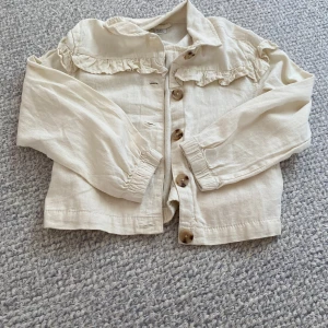 Beige overshirt med volang och knappar - Snygg och super söt beige overshirt i linneblandning med volangdetaljer över axlarna och stora knappar framtill. Jackan har långa ärmar med resår vid handlederna och en avslappnad passform. Perfekt för dig som gillar en trendig look. Så söt! Säljs för att jag tyvärr glömt bort att jag har den och den nu är för liten för mig. 