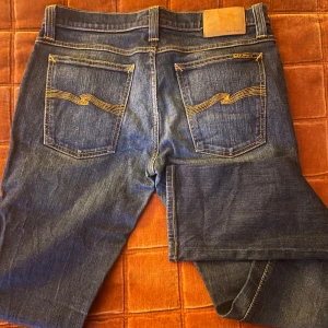 Mörkblå jeans från Nudie Jeans - Säljer ett par mörkblå jeans från Nudie Jeans med klassisk femficksdesign och snygga kontrastsömmar. Jeansen har raka ben och normal passform, med Nudie-loggan broderad på bakfickorna. Tillverkade i slitstarkt denim av bomull.