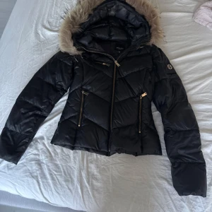 Svart pufferjacka från Hollies  - Säljer en svart pufferjacka från Hollies med snygg päls på huvan och guldiga dragkedjor. Jackan är quiltad och har två fickor framtill. Perfekt för kalla dagar och riktigt skön att ha på sig. 