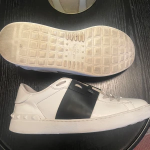 Valentino Open sneakers - Säljer ett par Valentino Open sneakers i vitt skinn med bred svart läderdetalj över sidan. Skorna har klassisk snörning, platt sula och coola nitar baktill för extra edge. Perfekta för dig som gillar stilrena men ändå unika sneakers. Lite slitna i sulan.