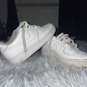 Vita Nike Air Force 1 sneakers - Klassiska vita Nike Air Force 1 sneakers med lågt skaft och snörning. Skorna har en rund tå, platt sula och är tillverkade i skinn med textildetaljer på insidan. Perfekta för dig som gillar streetstyle och vill ha en ikonisk sneaker i garderoben. Skon har dock några slitna ställen